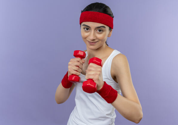 哑铃身穿运动服的年轻健身女士举着两个哑铃 像一个拳击手站在紫色的墙上运动装喜欢站