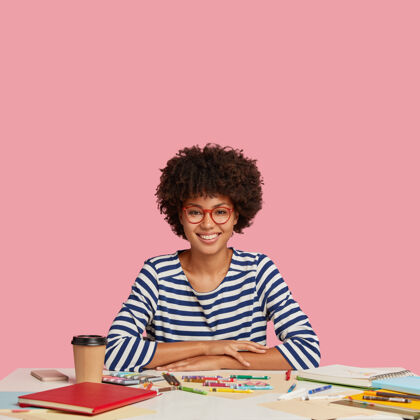 螺旋形快乐成功的设计师穿着水手毛衣 手放在桌子上 用蜡笔画杰作 笑容灿烂 享受外卖咖啡 隔离在粉红色的墙上 有自由的文字空间平面图女人蜡笔