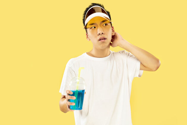 面部黄色工作室背景上的韩国年轻人半身像穿着白衬衫 戴着黄色帽子的男模喝鸡尾酒人类情感的概念 表达 夏天 假期 周末迪斯科舞蹈帽子