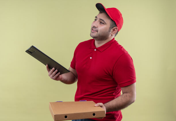 姿势年轻的送货员身穿红色制服 头戴鸭舌帽 手里拿着披萨盒和剪贴板 脸上带着微笑站在绿色的墙上 望着一旁帽子递送盒子