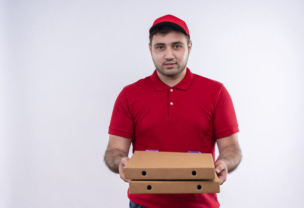 男人年轻的送货员身穿红色制服 头戴鸭舌帽 手里拿着披萨盒 站在白色的墙上自信地微笑着看着送货披萨