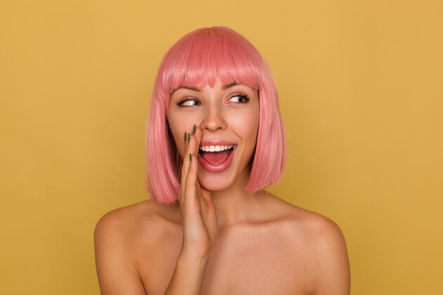 发型积极的年轻迷人的粉红色头发的女人 短发时髦 分享秘密 保持手掌靠近她的嘴 微笑着站在芥末墙站立发型爱情