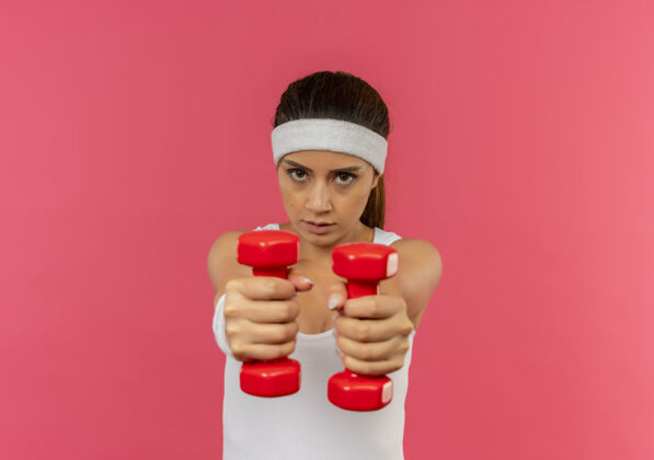 站着身穿运动服 头箍 举着两个哑铃 站在粉色墙壁上做运动的年轻健身女士 看上去很自信健康运动装健身