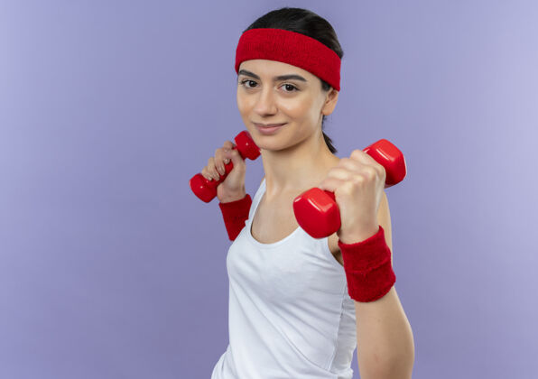 运动身穿运动服 头箍 双手举着两个哑铃 面带微笑 自信地站在紫色墙壁上锻炼身体的年轻健身女士自信手姿势