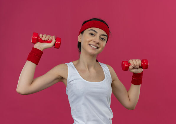 举起身穿运动服 头箍 双手举着两个哑铃 面带微笑 自信地站在粉色墙壁上锻炼身体的年轻健身女士运动装运动锻炼