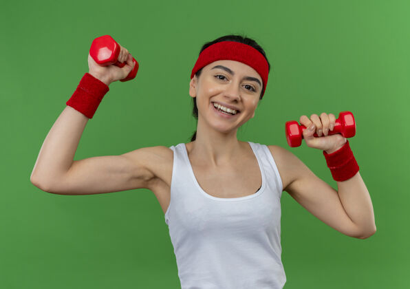 健身身穿运动服的年轻健身女士 头戴钢带 手持两个哑铃 举手练习 微笑自信地站在绿色的墙上自信运动装微笑