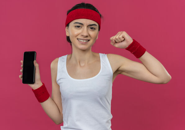 健身身穿运动服 头戴智能手机的年轻健身女士站在粉红色的墙上 握紧拳头 表情咄咄逼人站立侵略性显示