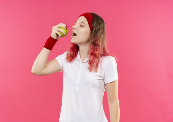 姿势戴着头巾的年轻的运动型女人举着绿色的苹果站在粉红色的墙上要咬它运动员女人苹果