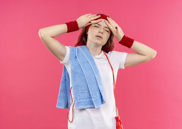 运动戴着头巾 肩上扛着毛巾 站在粉红色的墙上 看上去疲惫而疲惫的年轻运动女性运动装姿势运动员