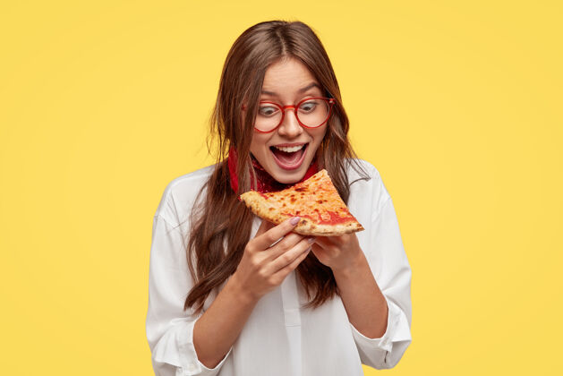 切片饥肠辘辘的学生一边张大嘴巴一边看美味的披萨片 一边想吃 穿着白衬衫 模特靠着黄色的墙壁积极的女人拿着垃圾食品人和吃惊呆了白种人年轻人