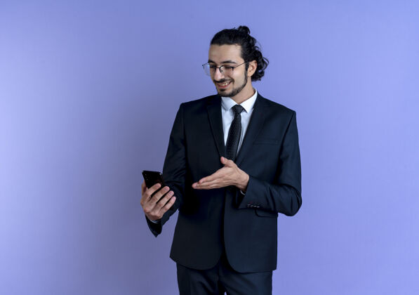 姿势一个穿着黑色西装戴着眼镜的商人站在蓝色的墙上看着智能手机的屏幕微笑着男士眼镜优雅