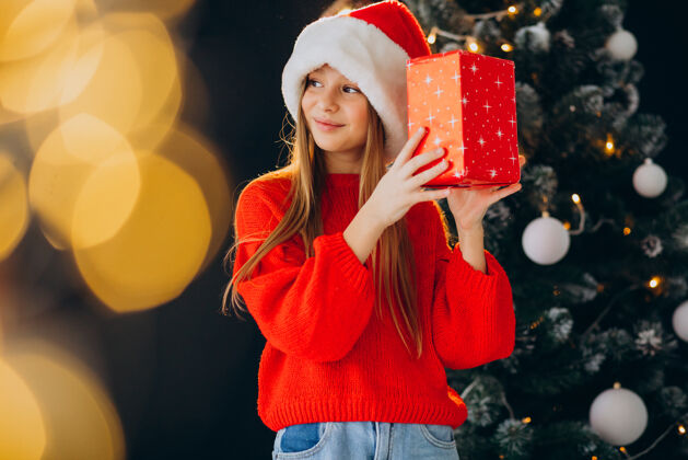 乐趣可爱的少女戴着圣诞树旁的红色圣诞帽看积极人