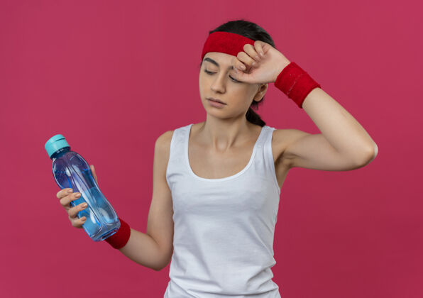 看穿着运动服的年轻健身女 头上戴着头巾 手里拿着一瓶水 站在粉红色的墙上 看上去很累健身市民姿势