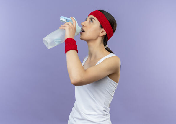 年轻穿着运动服的年轻健身女 头上戴着头巾 手里拿着一瓶水 站在紫色的墙上 看上去很累抱着水姿势