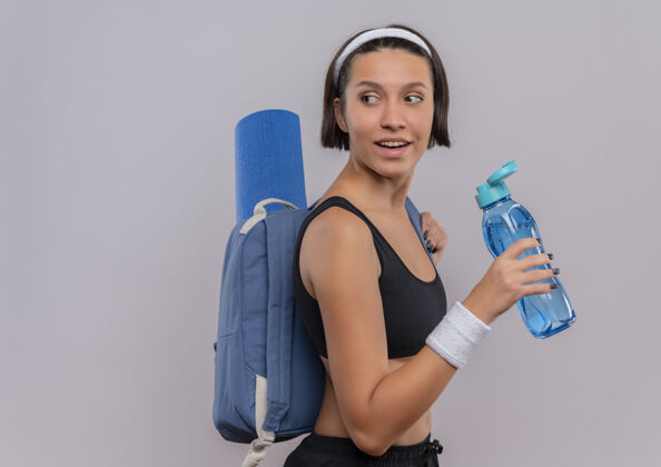 背包年轻的健身女士穿着运动装 背着背包和瑜伽垫 手里拿着一瓶水 面带微笑地站在白色的墙上 望着一旁瓶子水微笑
