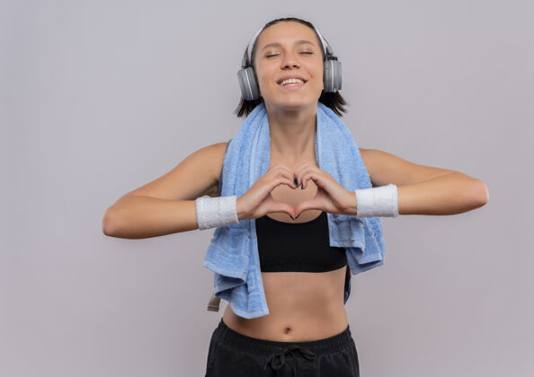 人身穿运动服的年轻健身女士头戴耳机 脖子上套着毛巾 闭着眼睛用手指做心脏手势 站在白墙上感受积极的情绪手指积极人