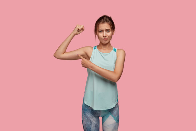 皱眉照片中的女运动员表情不悦 举手展示肌肉 表示在肱二头肌 穿着休闲t恤和紧身裤 隔离在粉红色的墙壁上训练理念不高兴运动员时尚