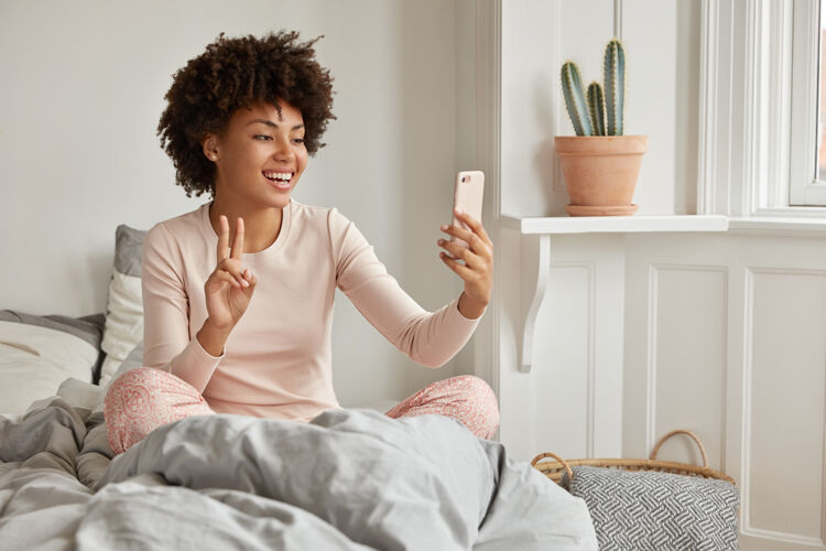 非洲室内拍摄的是一位穿着休闲睡衣的快乐深色皮肤的女士 有早晨的视频通话 手持现代手机 在床上摆姿势 面带笑容 在屏幕上显示和平标志早上自拍姿势视图放松