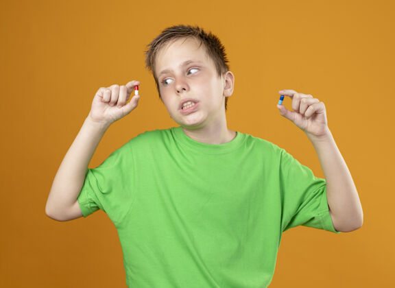 男孩生病的小男孩穿着绿色t恤 身体不适 手里拿着药片 站在橙色的背景上 困惑地试图做出选择站立药片感觉