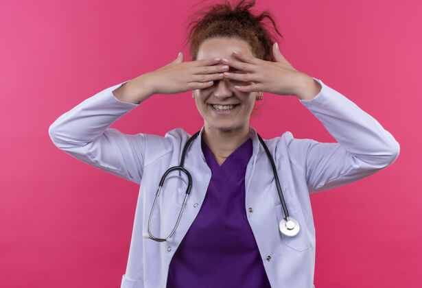 戴着年轻的女医生穿着白色外套 戴着听诊器 站在粉红色的墙上微笑着闭上眼睛微笑手指听诊器