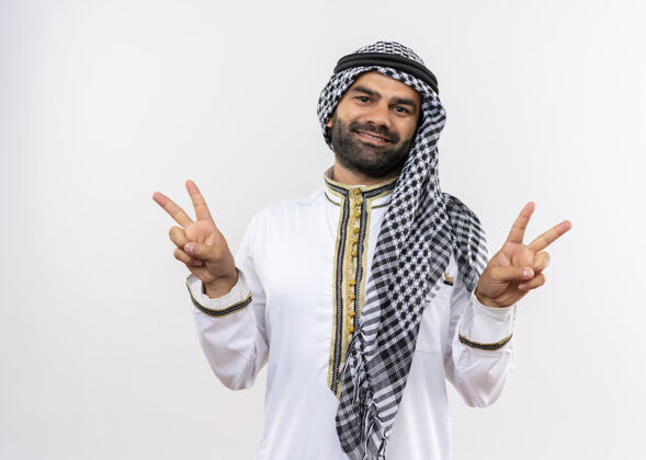 两者身着传统服装的阿拉伯男子微笑着积极快乐地展示着胜利的标志 双手站在白色的墙上阿拉伯语手展示