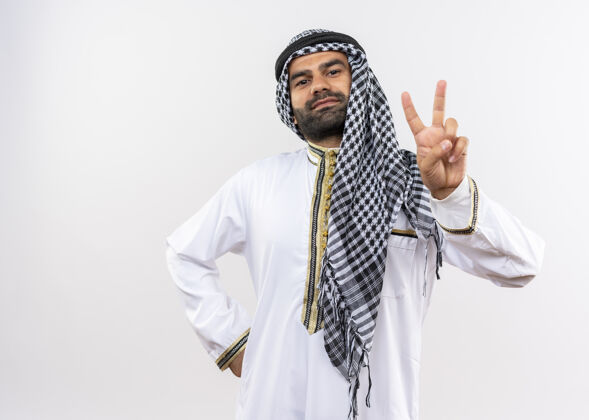 微笑身着传统服装的阿拉伯男子微笑自信地站在白墙上展示胜利标志展示自信站立