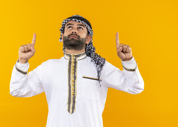 手指身着传统服装的阿拉伯商人抬起头 严肃地用手指指着橙色的墙壁认真穿看
