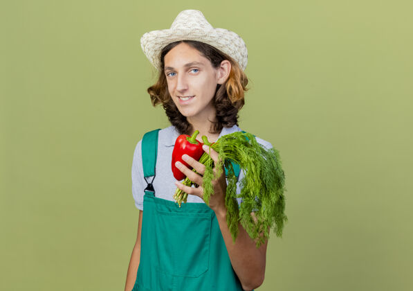 抱着年轻的园丁 穿着连体衣 戴着帽子 手里拿着红甜椒和新鲜的草药站着年轻微笑