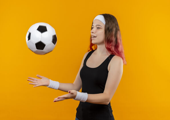 人穿着运动服的年轻健身女士快乐而积极地站在橙色的墙上扔足球市民健身积极