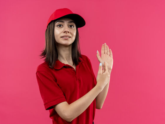 交货穿着红色马球衫和帽子的年轻送货女孩看着摄像机微笑着指着她的手臂 手指站在粉色的背景上衬衫指着年轻