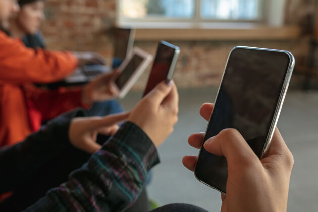 一起一群快乐的年轻人一起坐在沙发上分享智能手机上的新闻 照片或视频 阅读文章或玩游戏 享受乐趣社交媒体 现代科技移动男性女孩