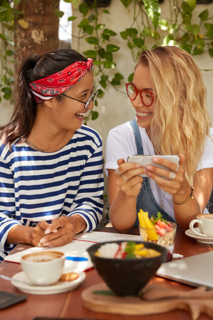 手机两个快乐的朋友在手机上看视频 课后喝咖啡休息 戴眼镜 吃美味的沙拉和喝咖啡 对着舒适的咖啡馆内部摆姿势 连接无线互联网团友甜点室内