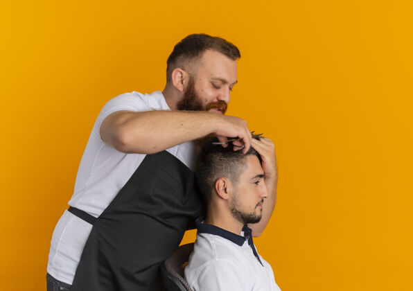做胡子专业的胡须理发师围着围裙用剃须刀机给站在橘色墙上的年轻人理发刮胡子理发师机器