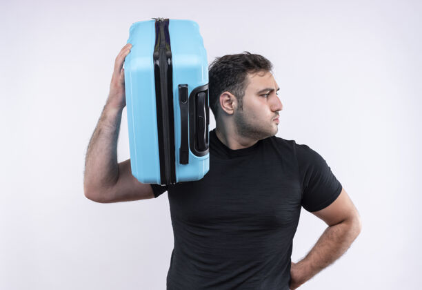 市民一个穿着黑色t恤的年轻旅行家拿着手提箱 严肃的脸站在白色的墙上望向一边旅行者夏天严肃