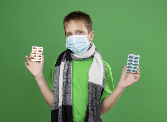 小生病的小男孩穿着绿色t恤 脖子上围着暖和的围巾 戴着护面面具 拿着药片 站在绿色背景上看着摄像机男孩疾病保护