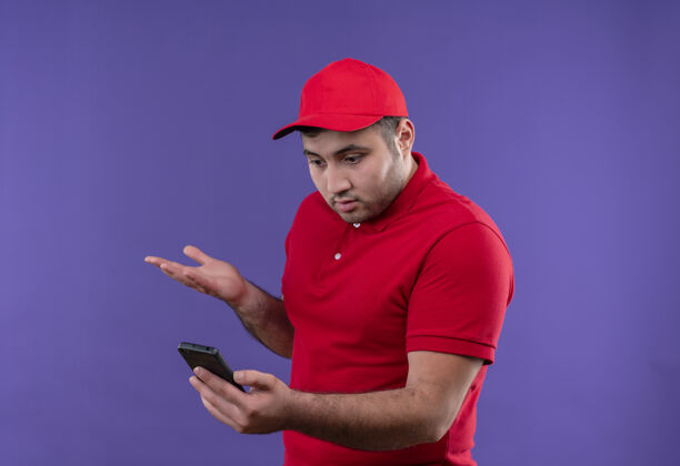 男人年轻的送货员穿着红色制服 戴着帽子 站在紫色的路边 困惑而失望地看着智能手机的屏幕公民制服姿势