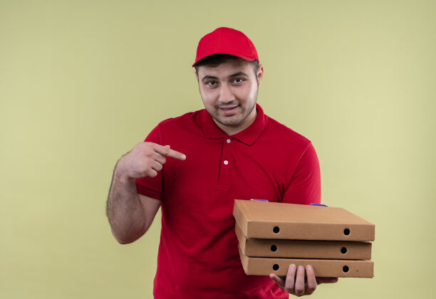 年轻人年轻的送货员身穿红色制服 头戴鸭舌帽 拿着披萨盒 手指着披萨盒 站在绿色的墙上自信地微笑着人披萨帽子