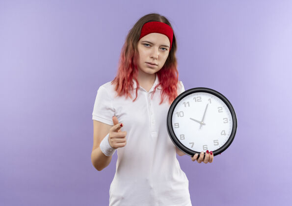 人年轻的运动女孩拿着挂钟严肃的脸指着手指站在紫色的墙上运动年轻人钟