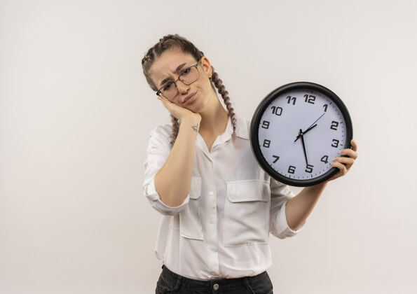 人戴着眼镜 梳着辫子 穿着白衬衫的年轻女学生手里拿着挂钟 站在白墙上看得又累又无聊眼镜辫子姿势