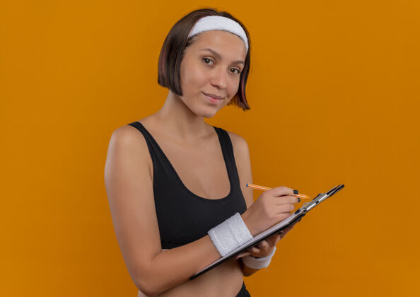 人身穿运动服的年轻健身女 头上戴着头巾 手里拿着写字板 空白的书页写着 站在橙色的墙上 看起来很自信公动员空白