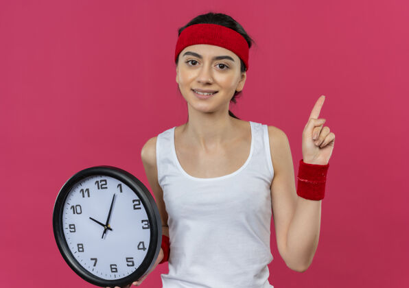 看身穿运动服的年轻健身女士 头上戴着手环 手持挂钟 食指自信地站在粉色的墙上钟墙表演