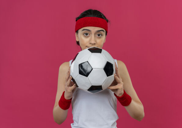 抱身穿运动服 头戴足球带的年轻健身女士站在粉红色的墙上看着摄像机 神情困惑足球人运动