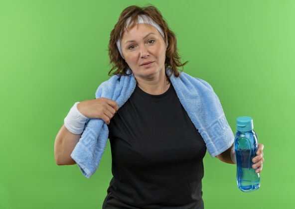 困惑身穿黑色t恤的中年运动女性 脖子上戴着头带和毛巾 手里拿着一瓶水站在绿色的墙上女人看头带