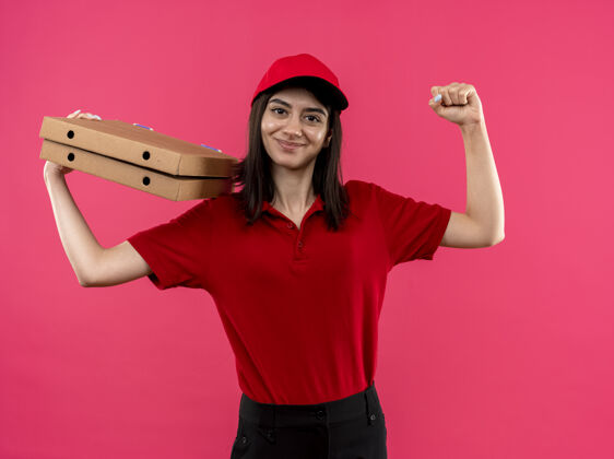 衬衫年轻的送货女孩穿着红色马球衫 戴着帽子 手里拿着比萨饼盒 举起拳头 开心而积极地微笑 友好地站在粉色背景下披萨举行盒子