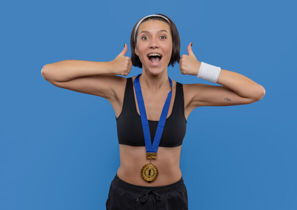 摆姿势一位身穿运动服 脖子上挂着金牌的年轻女子竖起大拇指 为自己的成功而欢欣鼓舞 她站在蓝色的墙上 快乐而兴奋脖子运动装展示