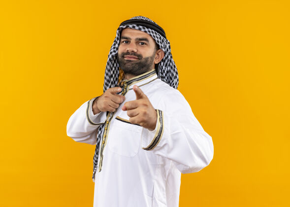 微笑身着传统服装的阿拉伯商人站在橙色的墙上手指着微笑自信地站着手指穿着站立