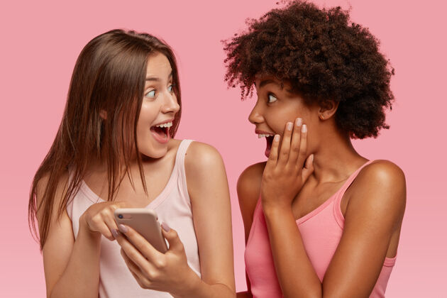 惊讶惊喜快乐的异族朋友开心地看着手机屏幕 读着不可思议的东西 一起对着粉色的墙壁摆姿势 在网上购物种族和科技放松消息Omg