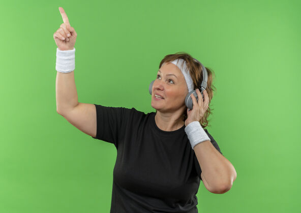 指向中年运动女性 身穿黑色t恤 戴着头带和耳机 看着一边 食指竖立在绿色的墙上旁边站耳机