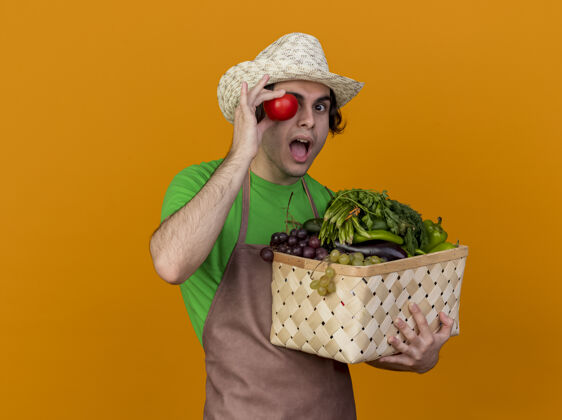 新鲜年轻的园丁 围着围裙 戴着帽子 手里拿着装满蔬菜和新鲜番茄的箱子 站在橙色的背景下微笑着 看上去很惊讶惊喜围裙举行