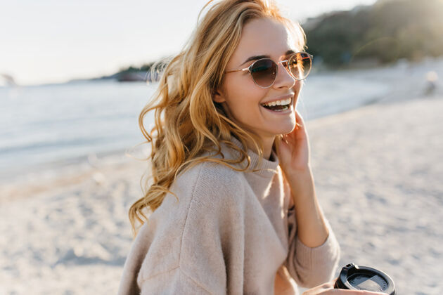 灵感可爱的女人真诚地笑着 在沙滩上放松戴着眼镜和毛衣的盲人女人拿着一杯咖啡天空乐趣海边